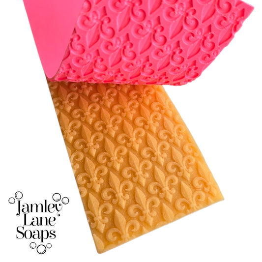 repeating fleur de lis pattern artisan polymer impression mat being peeled away from a gold melt and pour soap top.