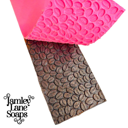a brown soap top showing the release of a coffee bean pattern created by the pink coffee bean mat thats shown partially peeled away.