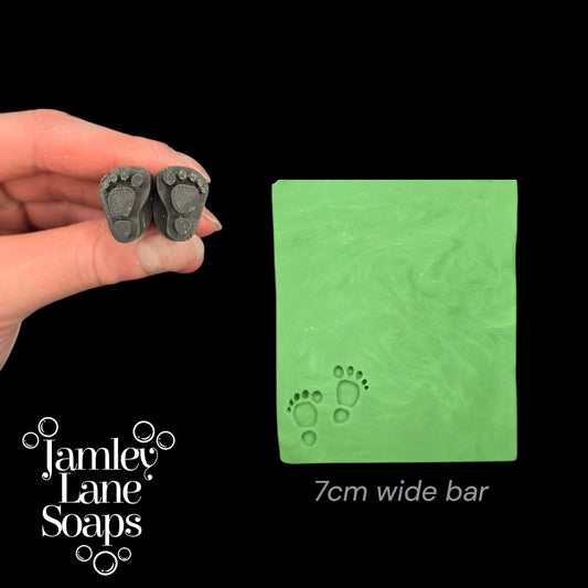 Hand holding two small gray footprints next to a green soap bar with footprints, branded as Jamley Lane Soaps.