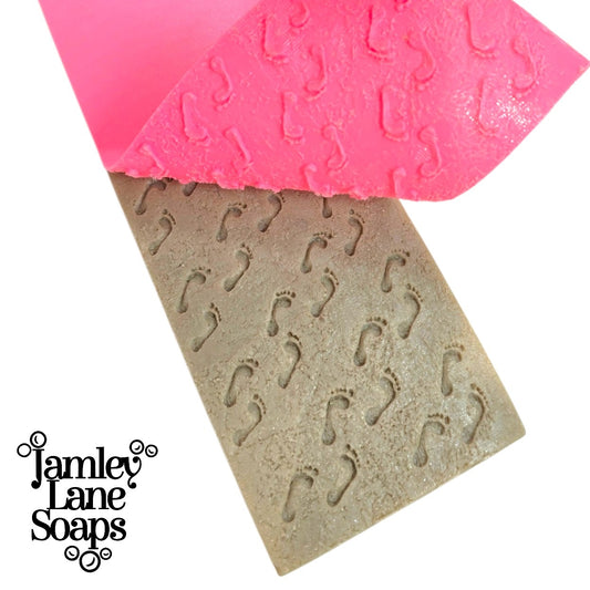 Pink artisan polymer impression mat being pulled away from  a sandy colored loaf of soap to reveal the pattern of footprints displayed with a jamley lane soaps logo overlay
